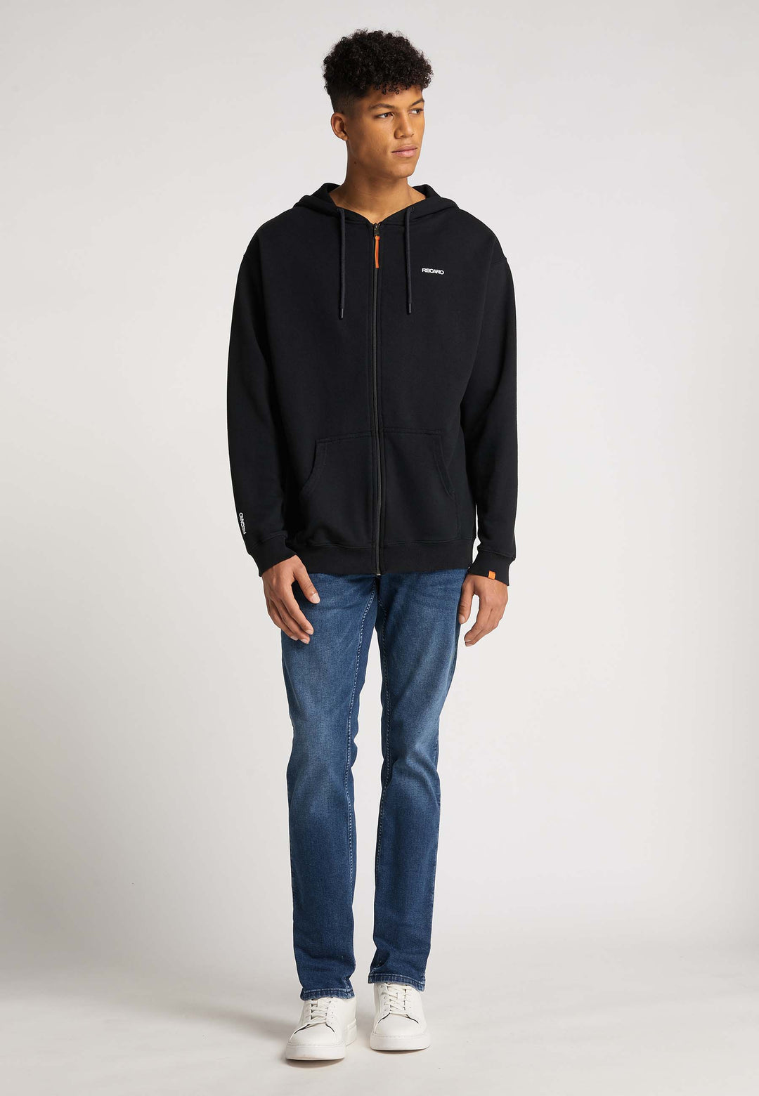 Ein junger Mann steht vor einem schlichten Hintergrund und trägt eine locker sitzende schwarze Hoodiejacke Originals von Recaro , dazu blaue Jeans und weiße Sneakers. Er blickt leicht zur Seite und hat entspannte Hände. Der Hoodie hat ein vielseitiges Unisex-Design.