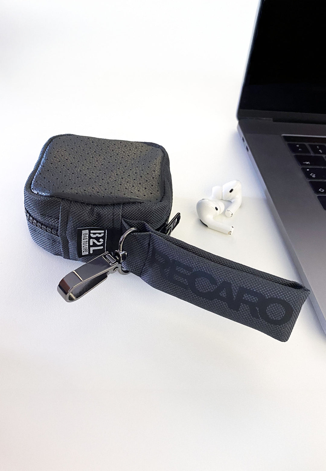 Das Tech & Travel Kopfhörer-Case aus dem RECARO Shop aus RECARO-Sitzleder inklusive Schlüsselanhänger und USB-Stick liegt neben Laptop und In-Ear-Kopfhörern auf einer weißen Fläche und demonstriert so stilvolles Upcycling.
