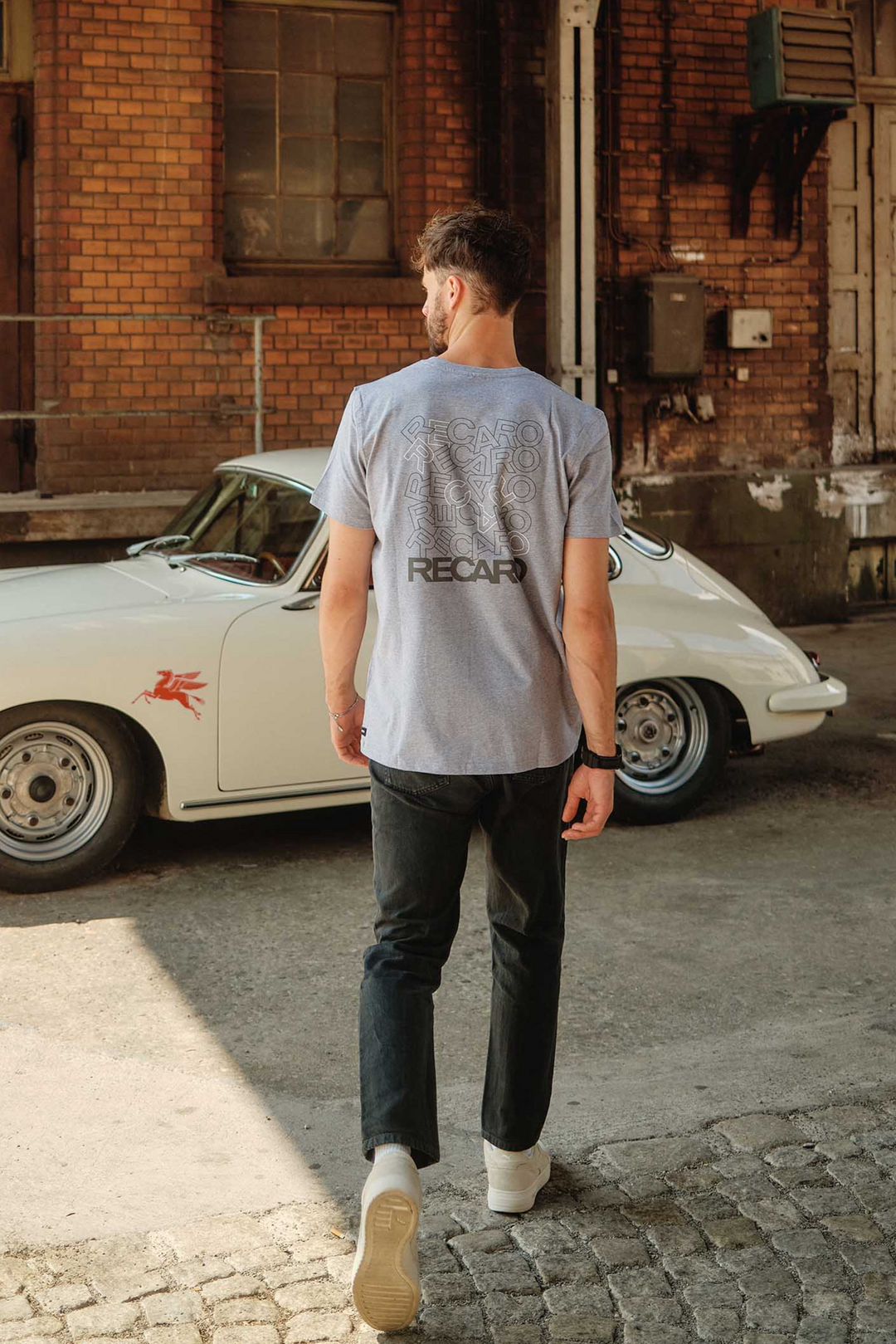 Ein Mann in einem grauen „RECARO“-T-Shirt mit Rückenaufdruck und schwarzen Jeans geht mit dem Rücken zur Kamera auf einen weißen Oldtimer mit roter Grafik und RECARO-Logo zu, der neben einem Backsteingebäude geparkt ist.