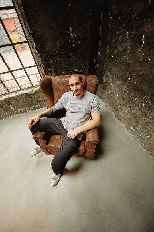 Ein Mann mit kurzen Haaren sitzt in einem braunen Sessel neben einem großen Industriefenster und trägt das RECARO T-Shirt Originals Embossed, dunkle Hosen und weiße Turnschuhe in einem Raum mit strukturierten dunklen Wänden und einem Betonboden.