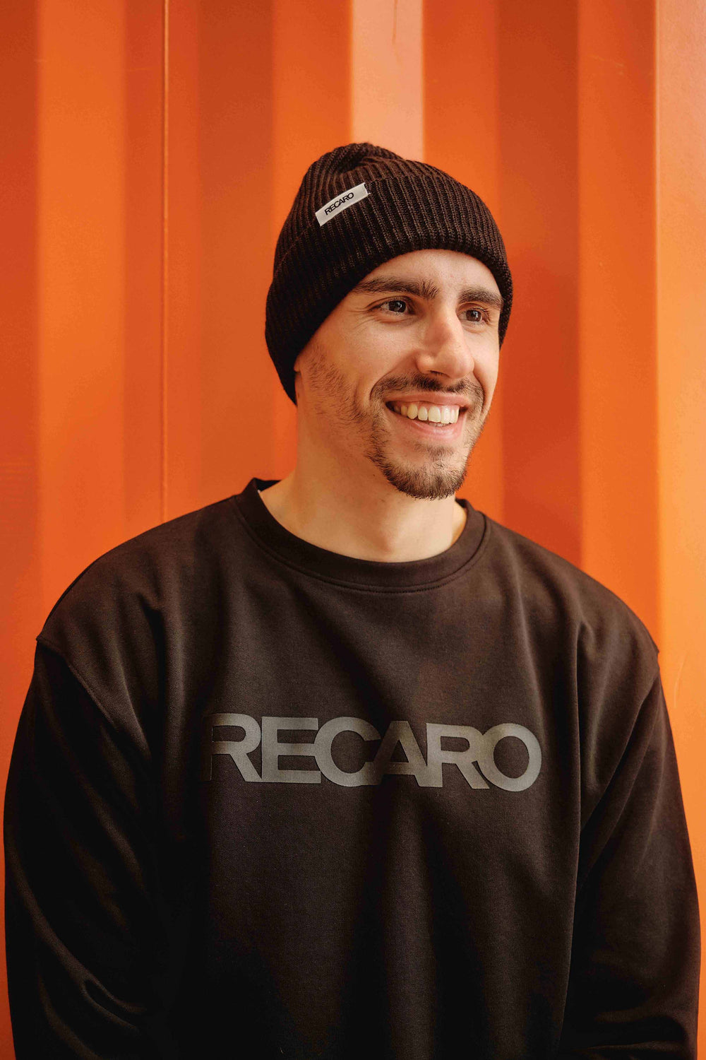 Ein lächelnder Mann mit kurzem Bart, der eine schwarze RECARO_Merchandising Beanie Originals und ein Sweatshirt aus Merinowolle trägt, steht für einen urbanen Look vor einer leuchtend orangefarbenen, geriffelten Wand.
