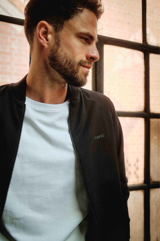 Ein bärtiger Mann mit kurzen Haaren, der einen RECARO_Merchandising Sweat Blouson über einem weißen T-Shirt trägt, steht drinnen an einem großen Fenster mit schwarzem Rahmen und blickt mit nachdenklichem Gesichtsausdruck zur Seite.