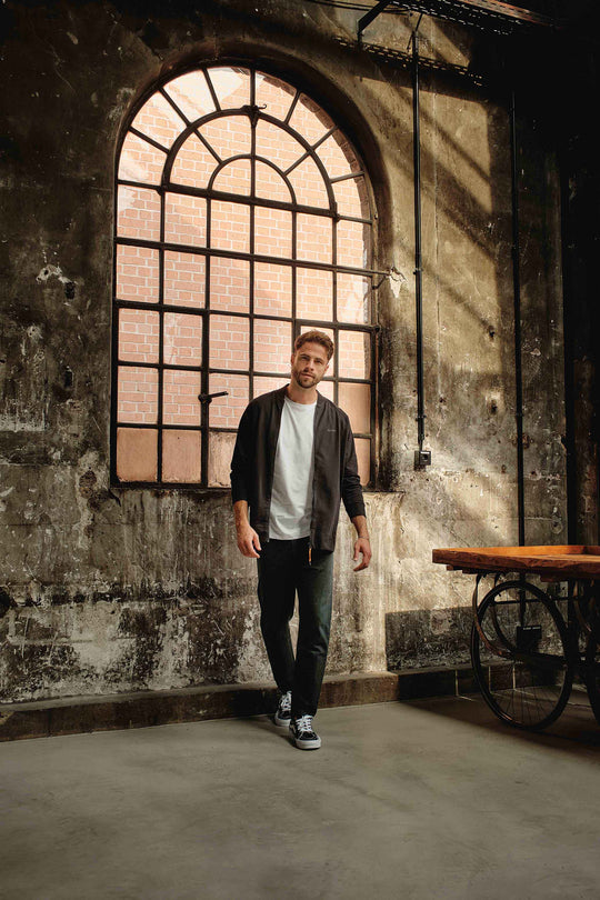 Ein Mann in Freizeitkleidung, gekleidet in einen RECARO_Merchandising Sweat Blouson mit Raglanärmeln und weichem Baumwollstoff, geht durch ein großes Bogenfenster ins Haus, während Sonnenlicht Schatten auf strukturierte Wände und einen mobilen Holztisch wirft.