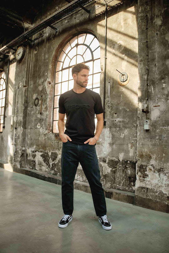 Ein Mann, der das RECARO T-Shirt Originals Embossed, dunkle Jeans und schwarze Turnschuhe trägt, steht mit den Händen in den Taschen vor großen Bogenfenstern in einem industriellen, verwitterten Gebäude mit strukturierten Wänden.