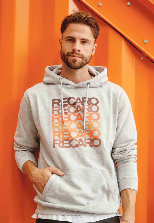 Ein Mann mit kurzen braunen Haaren und Bart trägt den hellgrauen RECARO Hoodie Spektrum mit RECARO-Aufdruck im Retro-Farbverlauf und steht vor einer orangefarbenen Metallwand.