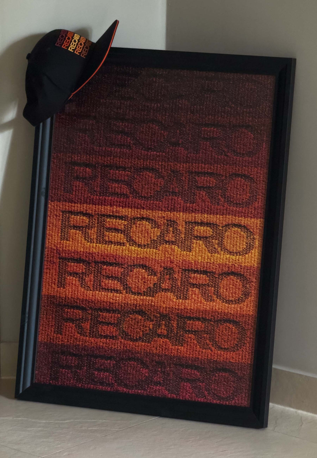 Ein schwarz gerahmtes RECARO Shop-Poster Spektrum 50 x 70cm mit rotem, orangefarbenem und gelbem RECARO Schriftzug lehnt an einer Wand, gekrönt in der linken Ecke von einer schwarzen Kappe mit orangefarbenem RECARO Branding als 1960er Jahre Wanddeko.