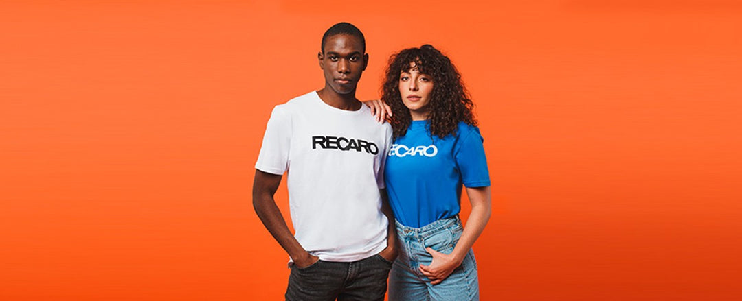 Vor einem orangefarbenen Hintergrund stehen zwei Personen. Eine trägt ein weißes RECARO T-Shirt, die andere ein blaues RECARO T-Shirt. Die Frau hat ihren Arm auf der Schulter des Mannes abgelegt.