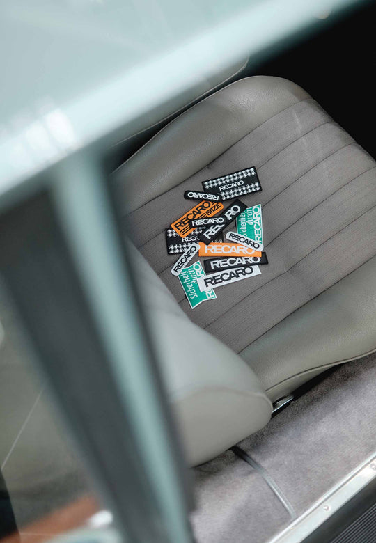 Ein grauer Autositz weist mehrere bunte RECARO Patch 110x25mm Logos von RECARO auf seinem strapazierfähigen Material auf, von oben durch das Fenster gesehen.