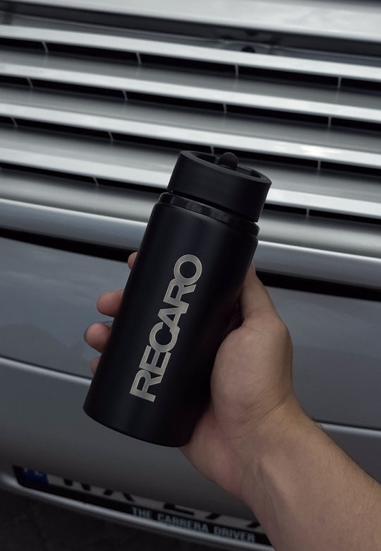 Eine Hand hält eine schwarze RECARO Shop Trinkflasche Edelstahl vor einem silbernen Auto mit horizontalen Kühlergrill-Lamellen im Hintergrund.