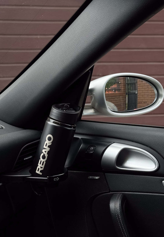 Eine schwarze RECARO Shop Trinkflasche Edelstahl im sportlichen Design sitzt in einem Becherhalter auf der Türverkleidung der Fahrerseite, durch das Fenster sind der Seitenspiegel und ein braunes Garagentor zu sehen.