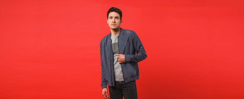 Vor einem schlichten roten Hintergrund steht ein Mann. Er trägt ein graues T-Shirt, dunkle Jeans und eine blaue Jacke und hält mit einer Hand den Reißverschluss der Jacke.