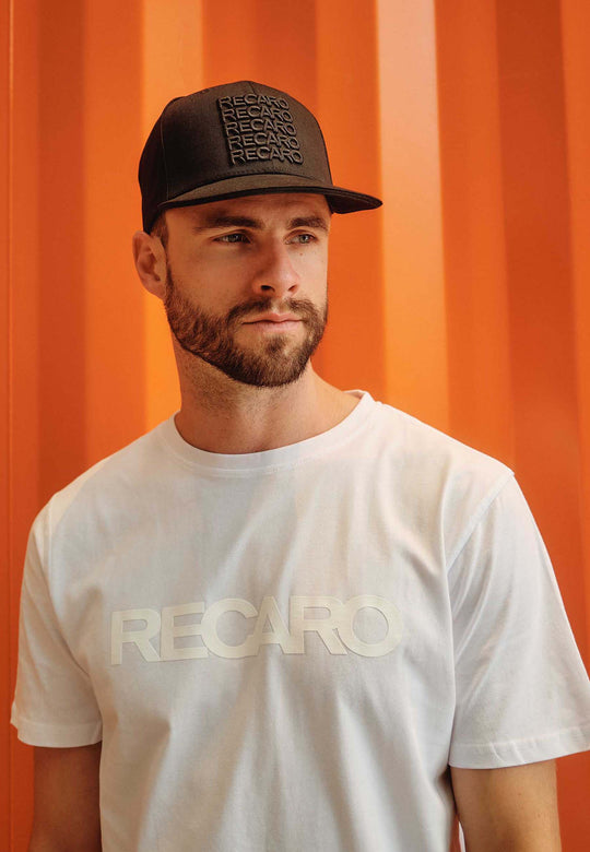 Ein bärtiger Mann mit einer schwarzen Snapback Cap Spektrum von RECARO_Merchandising und einem weißen RECARO T-Shirt steht vor einem orangefarbenen Hintergrund, sein Retro-Look wird betont, während er zur Seite blickt.