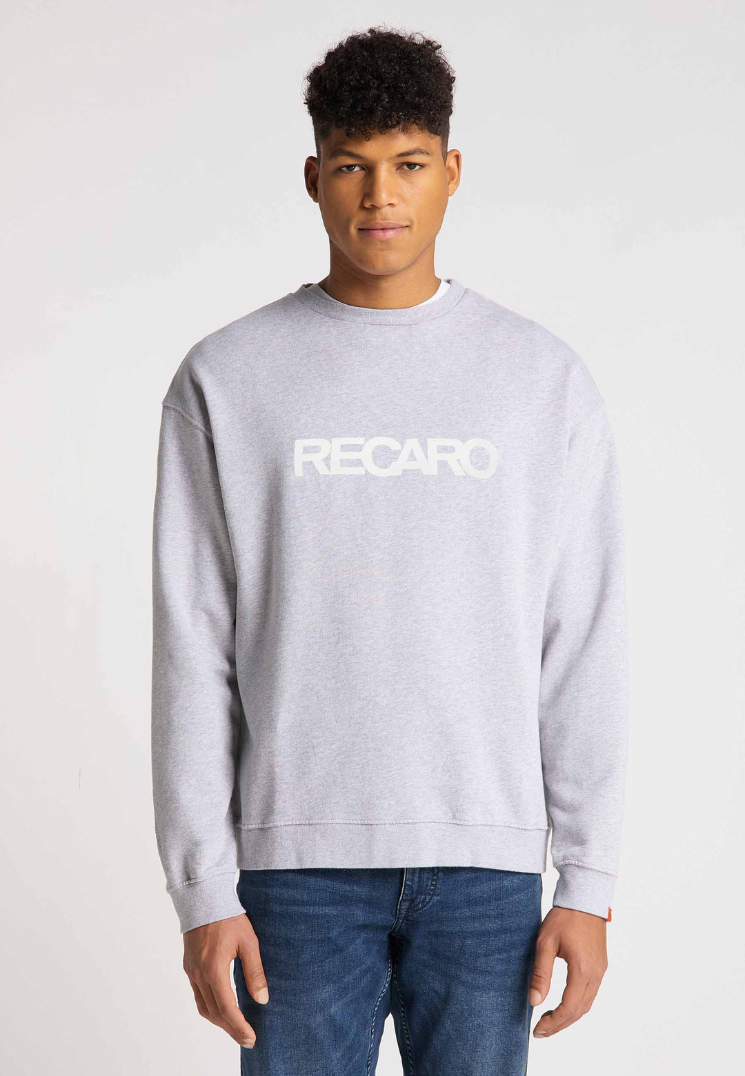Ein junger Mann mit kurzen lockigen Haaren trägt ein lässig geschnittenes RECARO Sweatshirt Originals Rubber Logo aus weicher Baumwollmischung mit weißem Schriftzug auf der Vorderseite, dazu Bluejeans auf schlichtem hellem Untergrund.
