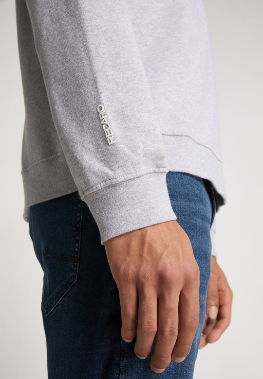 Eine Person trägt ein hellgraues RECARO Sweatshirt mit Originals Rubber Logo im Relaxed-Fit aus weicher Baumwollmischung und Bluejeans, seitlich abgebildet. Die Hand liegt entspannt neben ihr, am Handgelenk ist PE4GRO in Weiß gestickt.