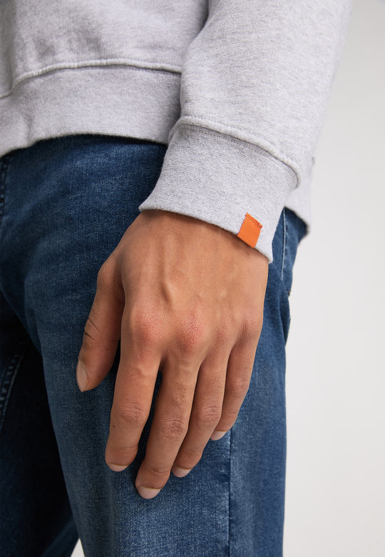 Eine Nahaufnahme einer Hand, die auf einer dunklen Jeans ruht. Sie trägt das RECARO Sweatshirt Originals Rubber Logo in Hellgrau, entspannter Schnitt, mit einem kleinen orangefarbenen Ärmeletikett und aus weichem Baumwollmischgewebe.