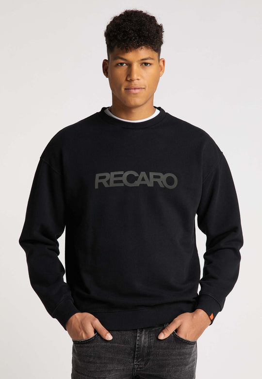 Ein junger Mann mit kurzen lockigen Haaren trägt ein lässig geschnittenes RECARO  Sweatshirt Originals Rubber Logo aus weichem Baumwollmischgewebe und steht mit den Händen in den Taschen vor einem schlichten hellen Hintergrund.