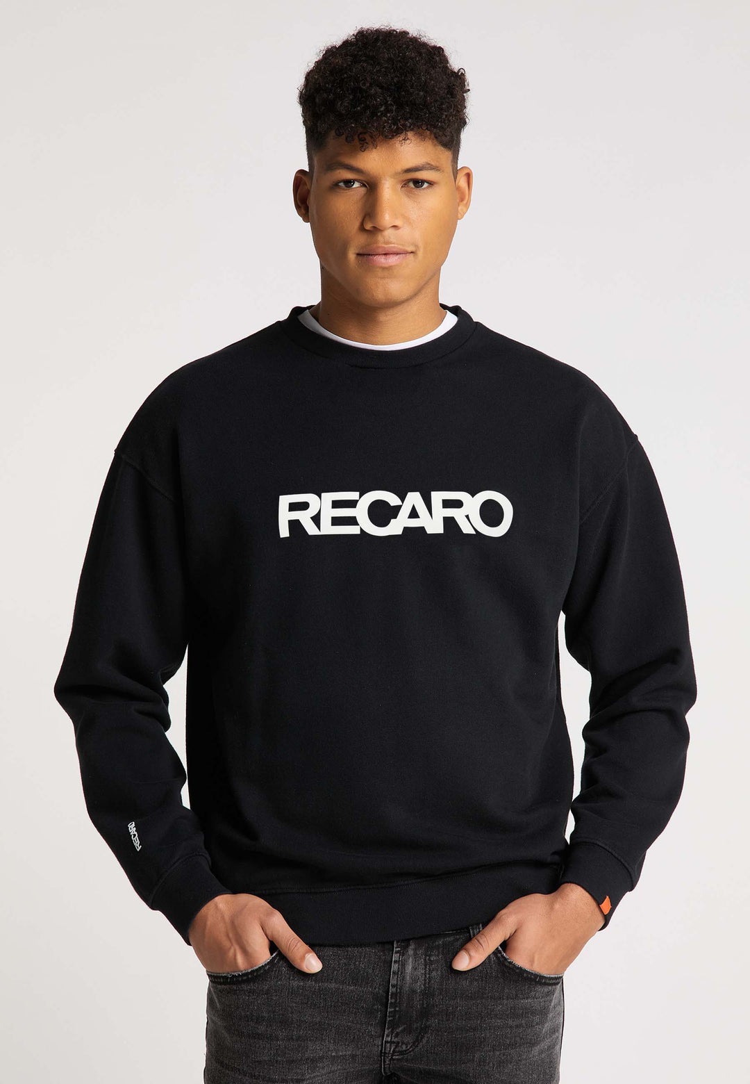 Eine Person steht mit Blick nach vorne und trägt ein Relaxed-Fit RECARO  Sweatshirt Originals Rubber Logo aus weicher Baumwollmischung mit dunklen Jeans vor einem schlichten hellen Hintergrund.