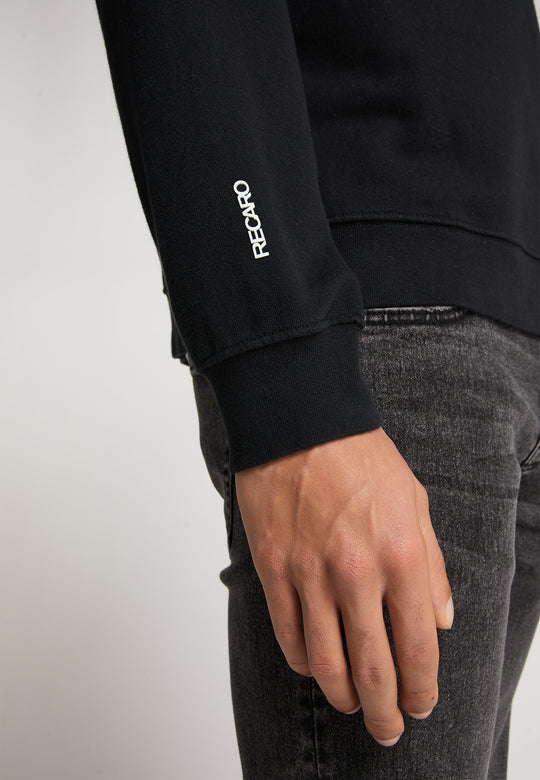 Nahaufnahme der rechten Hand einer Person, die ein schwarzes RECARO  Sweatshirt Originals Rubber Logo mit RECARO-Stickerei auf dem Ärmel trägt, kombiniert mit grauen Relaxed-Fit-Jeans. Der Hintergrund ist schlicht und hell.