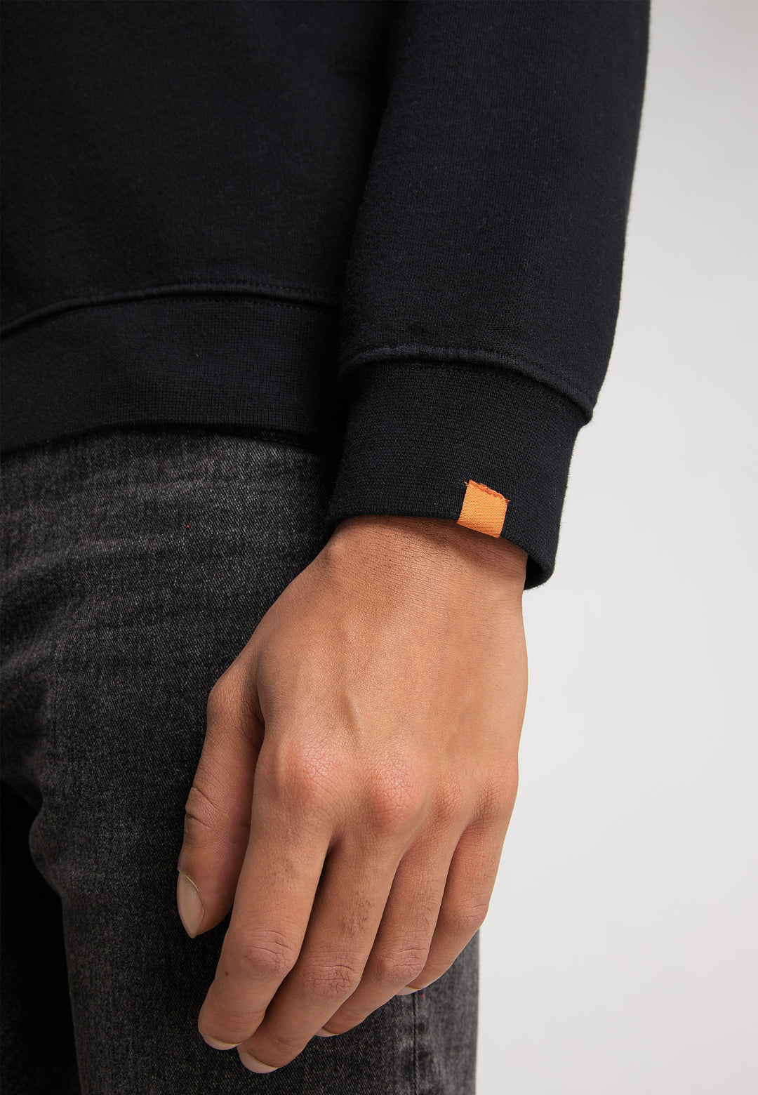 Nahaufnahme der linken Hand und des Handgelenks einer Person. Sie trägt ein schwarzes RECARO Sweatshirt Originals Rubber Logo mit orangefarbenem Manschettenetikett und eine locker sitzende dunkelgraue Jeans. Der Hintergrund ist schlicht und hell.