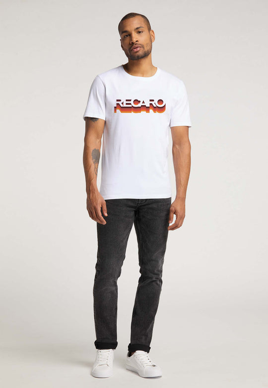 Ein Mann mit kurzen Haaren, Bart und einem sichtbaren Tattoo auf dem linken Arm präsentiert das RECARO T-Shirt Gradient mit Logo im Retro-Stil. Er trägt dazu schwarze Jeans und weiße Sneakers auf schlichtem Hintergrund.