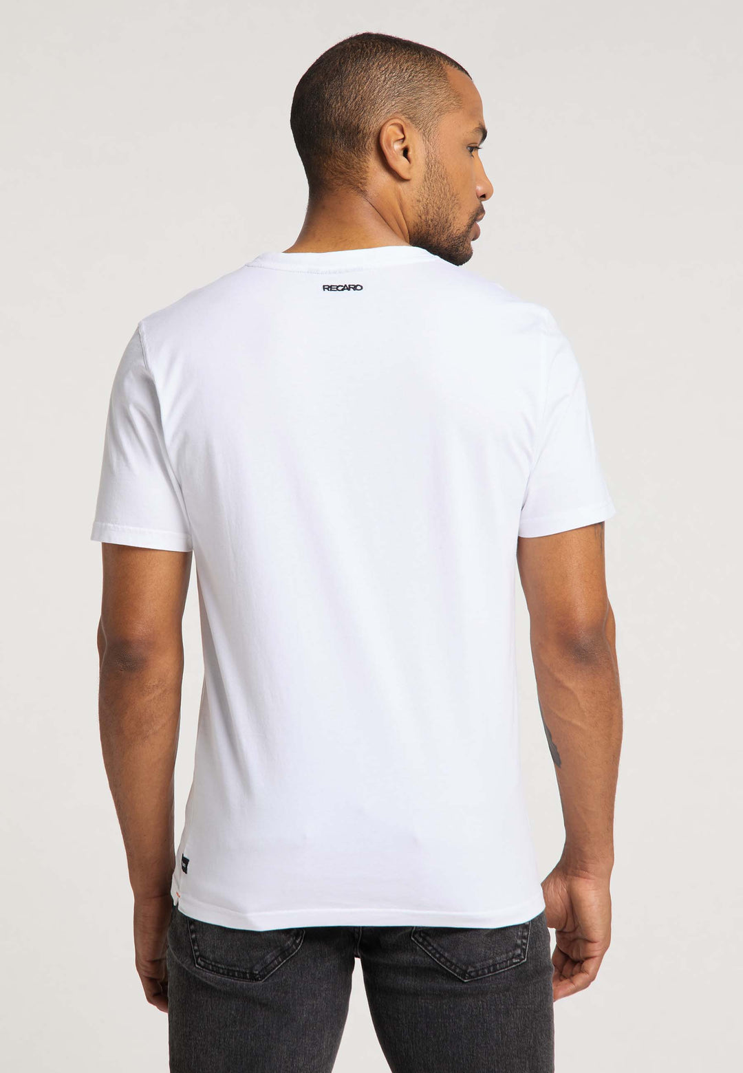 Ein Mann blickt von der Kamera abgewandt in einem schlichten weißen RECARO T-Shirt Gradient und dunkler Jeans. Ein kleines Logo am Rückenausschnitt hebt den dezenten Farbverlauf des Shirts vor hellem Hintergrund hervor.