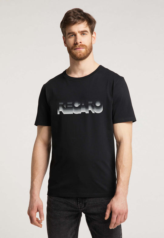 Ein hellhäutiger, braunhaariger Mann mit Bart trägt das RECARO T-Shirt Gradient – schwarz mit silberglänzendem RECARO Logo und dezentem Farbverlauf – und steht vor einem schlichten hellen Hintergrund, den Blick leicht an der Kamera vorbei gerichtet.