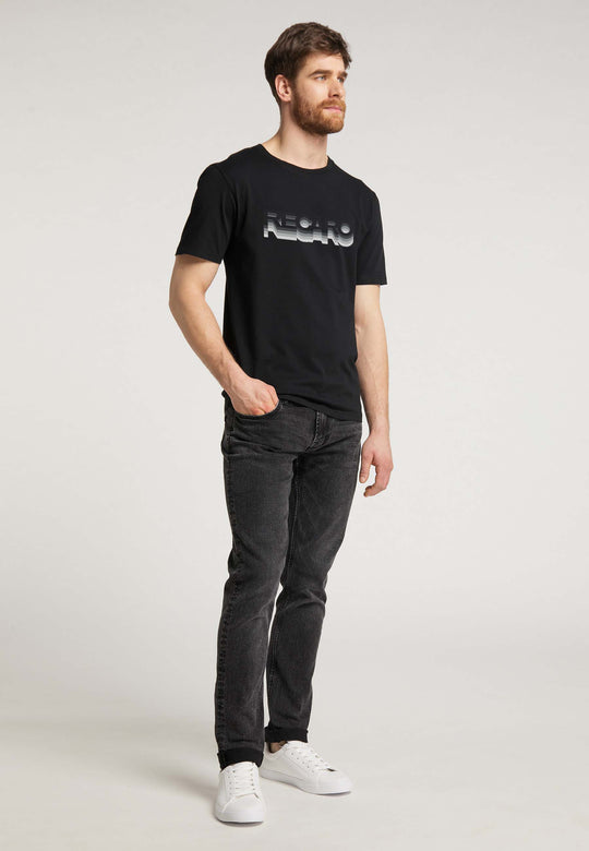 Ein bärtiger Mann steht vor einem schlichten Hintergrund. Er trägt das RECARO T-Shirt Gradient, dazu schwarze Jeans und weiße Sneakers. Eine Hand steckt in der Hosentasche, die andere ruht an seiner Seite.