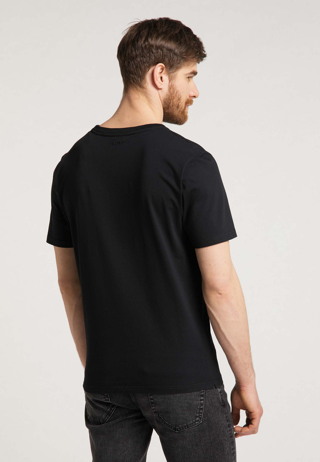 Ein Mann mit kurzen braunen Haaren und Bart blickt von der Kamera weg und trägt das RECARO T-Shirt Gradient und dunkle Jeans vor einem hellen, neutralen Hintergrund.