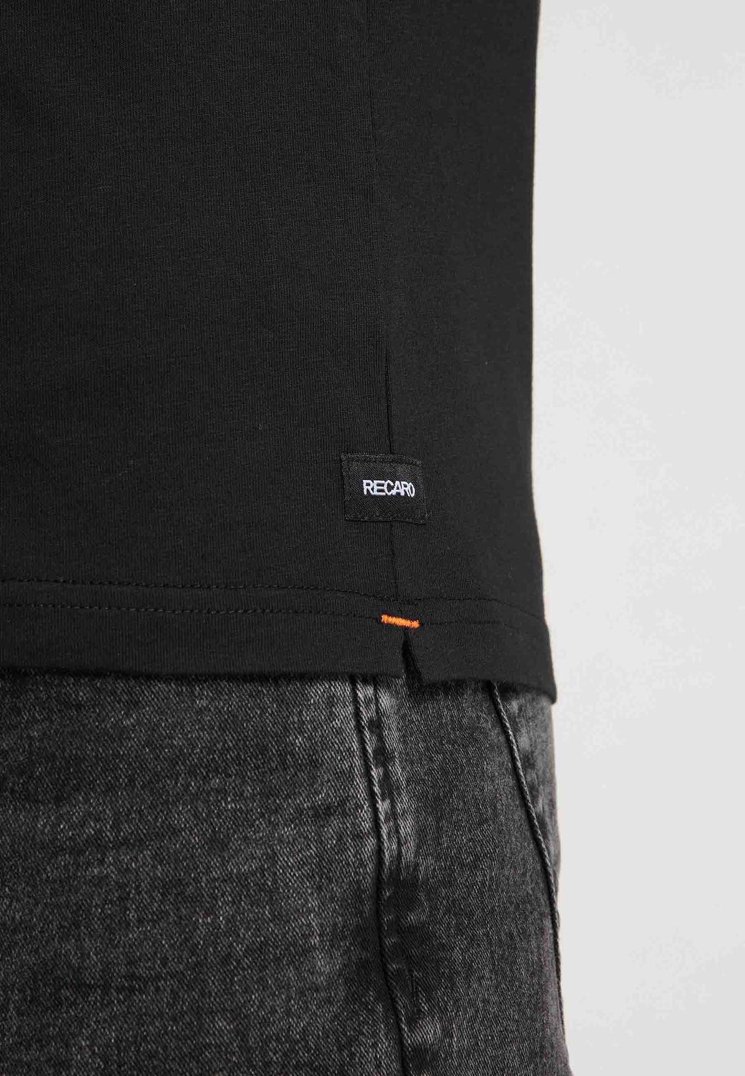 Nahaufnahme einer Person, die das RECARO T-Shirt Gradient in Schwarz mit einem kleinen schwarzen RECARO-Label am Saum trägt, kombiniert mit schwarzen Jeans vor einem schlichten hellen Hintergrund.