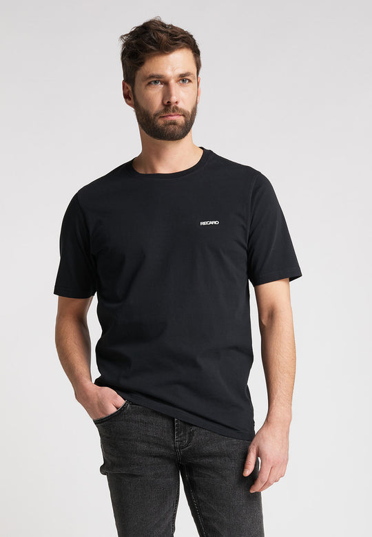 Ein Mann mit kurzen braunen Haaren und Bart trägt das Recaro_Merch_Daniel T-Shirt Originals Basic in Schwarz und dunkler Jeans und steht mit einer Hand in der Tasche vor einem schlichten hellen Hintergrund.