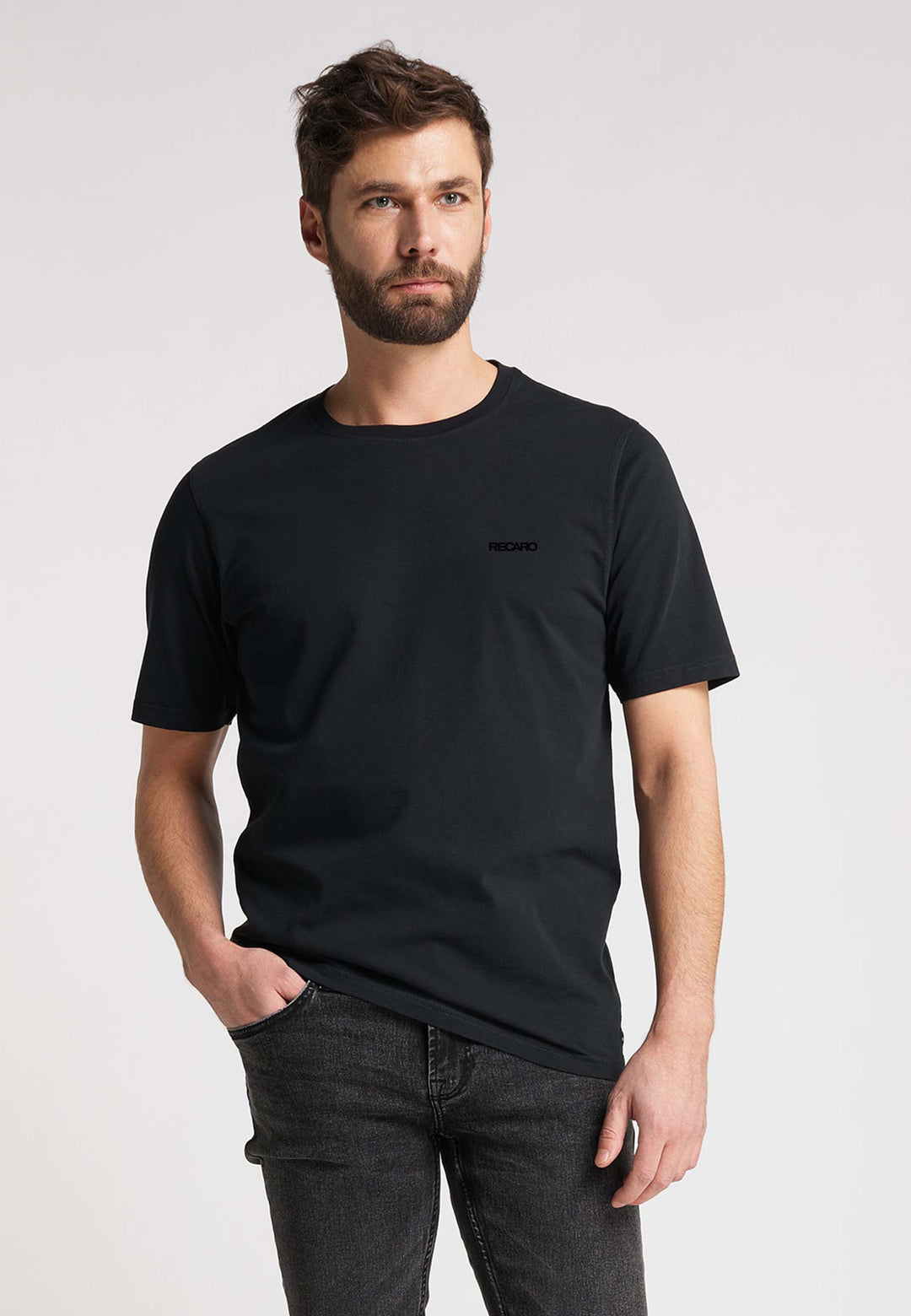 Ein Mann mit kurzen braunen Haaren und Bart trägt das Recaro T-Shirt Originals Basic in Schwarz und dunkler Jeans und steht mit einer Hand in der Tasche vor einem schlichten, hellen Hintergrund.
