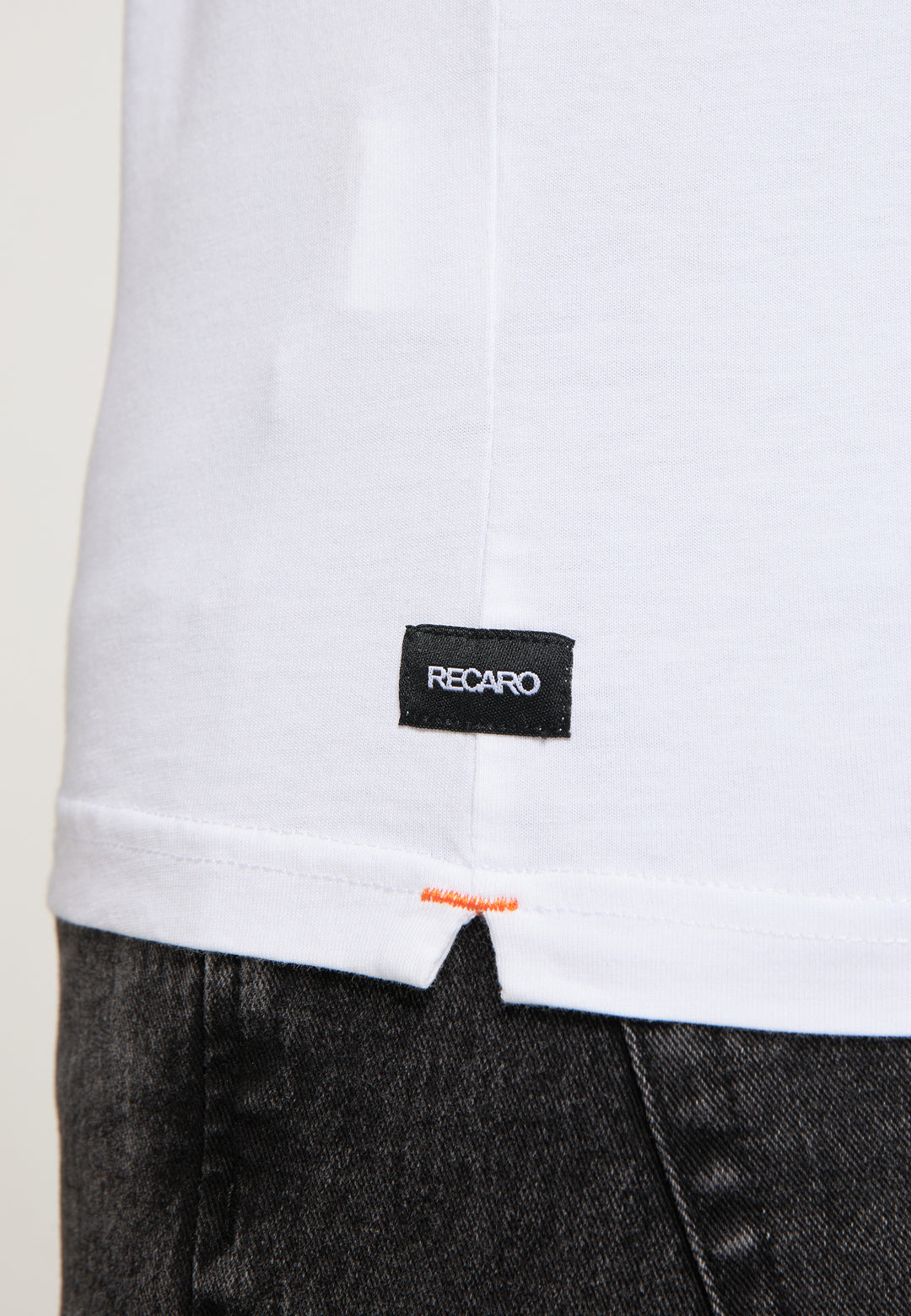 Nahaufnahme des unteren Teils des weißen T-Shirts Originals RECARO von Recaro_Merch_Daniel, Regular-Fit-Baumwolle, mit einem kleinen schwarzen RECARO-Etikett in der Nähe des Saums, getragen über dunkelgrauen Jeans.