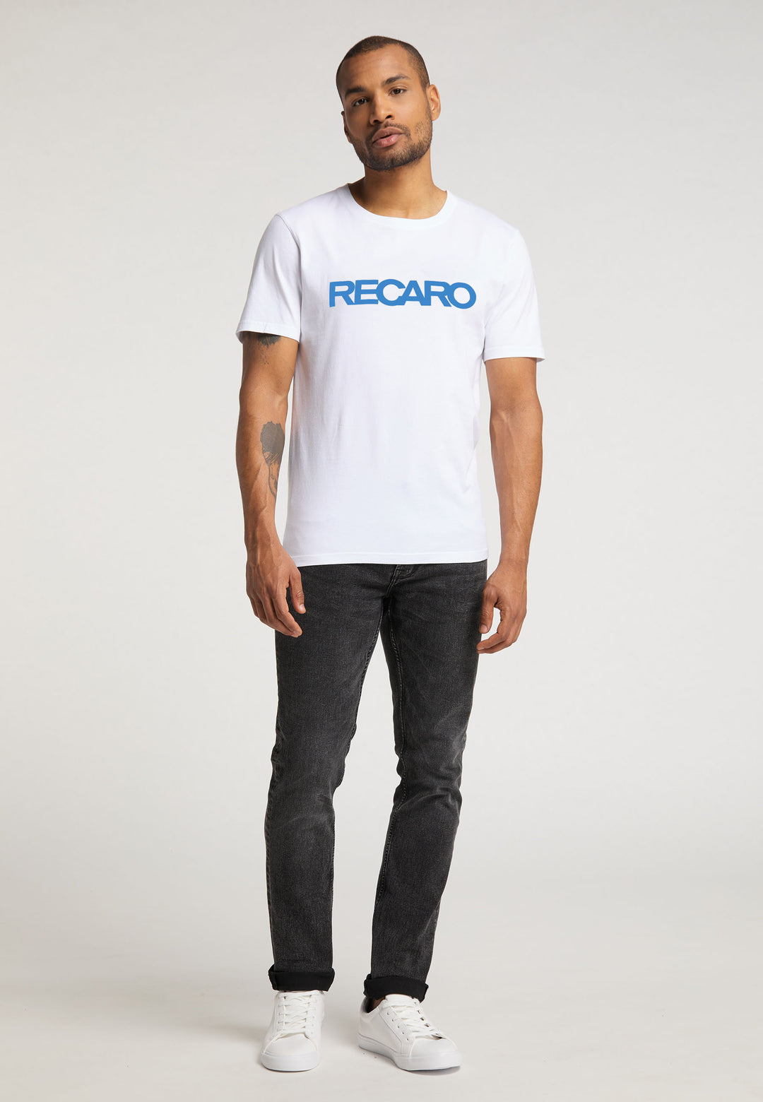Vor einem schlichten Hintergrund steht ein Mann mit kurzem Bart und Tattoo auf dem linken Unterarm. Er trägt ein weißes T-Shirt Originals RECARO von Recaro_Merch_Daniel mit blauer Schrift, schwarze Jeans und weiße Sneakers.