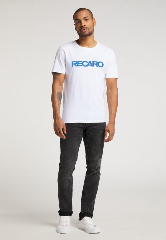 Vor einem schlichten Hintergrund steht ein Mann mit kurzem Bart und Tattoo auf dem linken Unterarm. Er trägt ein weißes T-Shirt Originals RECARO von Recaro_Merch_Daniel mit blauer Schrift, schwarze Jeans und weiße Sneakers.