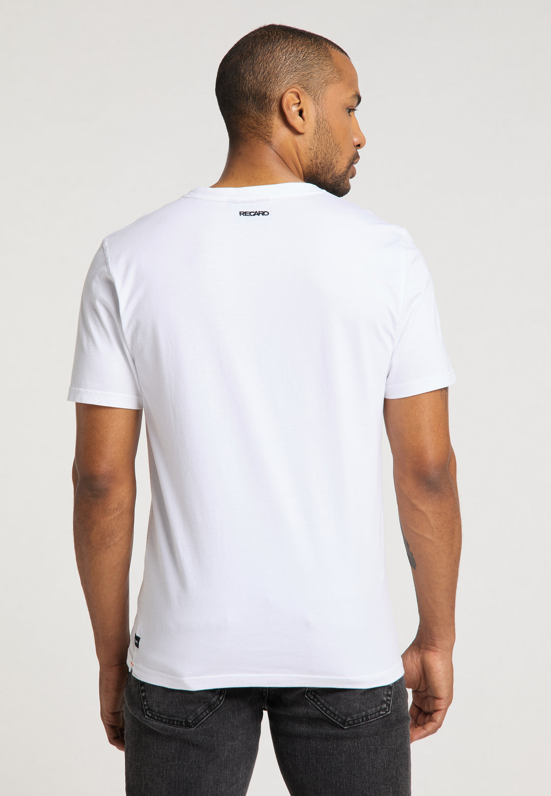 Ein Mann steht mit dem Rücken zur Kamera und trägt ein schlichtes weißes T-Shirt Originals RECARO von Recaro_Merch_Daniel und dunkle Jeans. Das Shirt hat ein kleines schwarzes Logo auf dem oberen Rücken vor hellem Hintergrund.