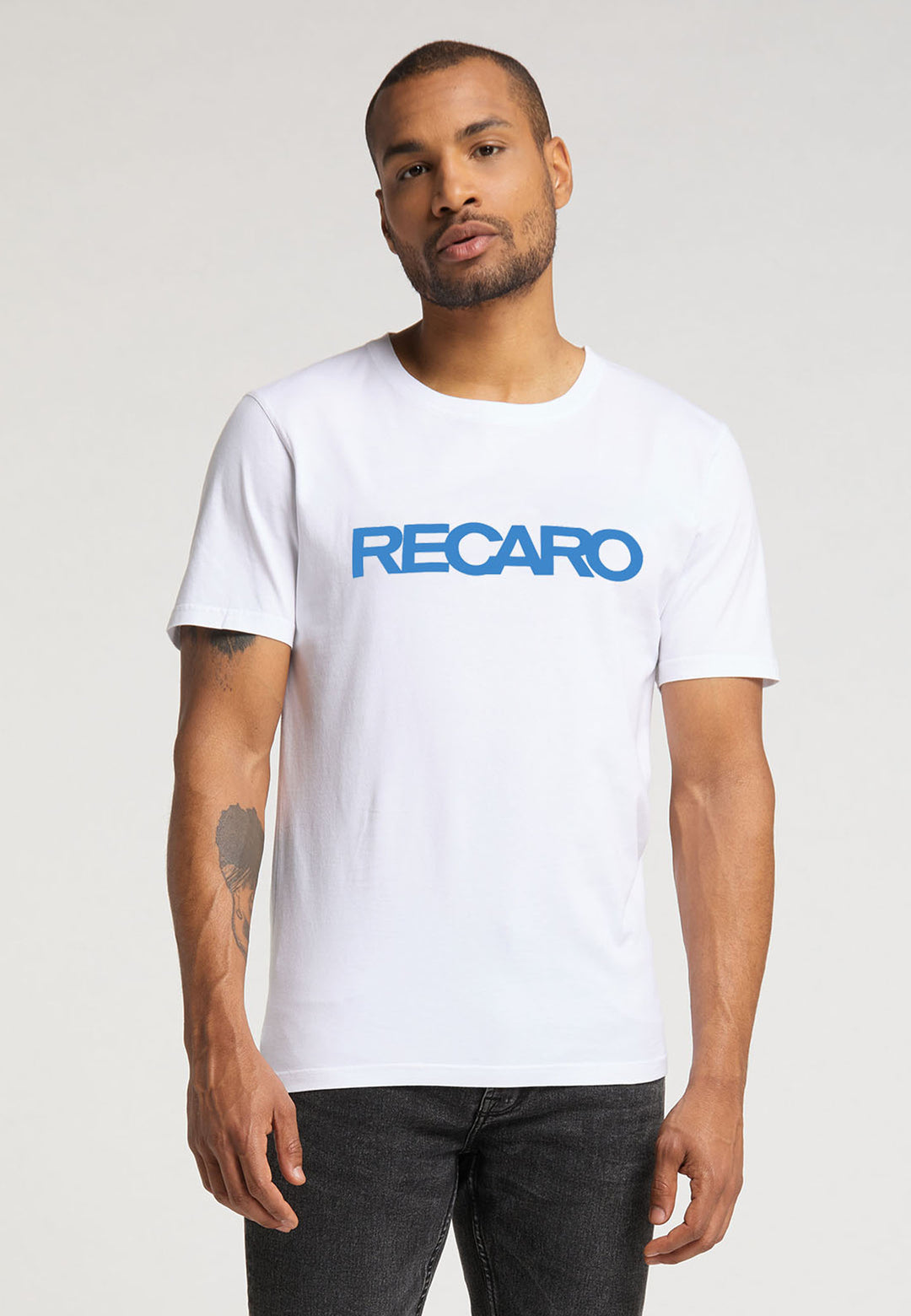 Ein bärtiger Mann trägt ein T-Shirt mit normaler Passform „Originals RECARO“ von Recaro_Merch_Daniel in Weiß mit blauer Schrift. Er steht vor einem schlichten Hintergrund, sein rechter Arm ist entspannt und auf seinem linken Unterarm ist ein Tattoo sichtbar.