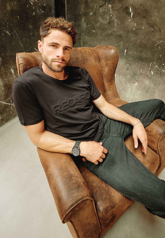 Ein Mann mit kurzen braunen Haaren und Bart sitzt entspannt in einem braunen Ledersessel, trägt ein schwarzes RECARO T-Shirt Originals Embossed, eine schwarze Hose und eine Uhr vor dem Hintergrund einer Betonwand.
