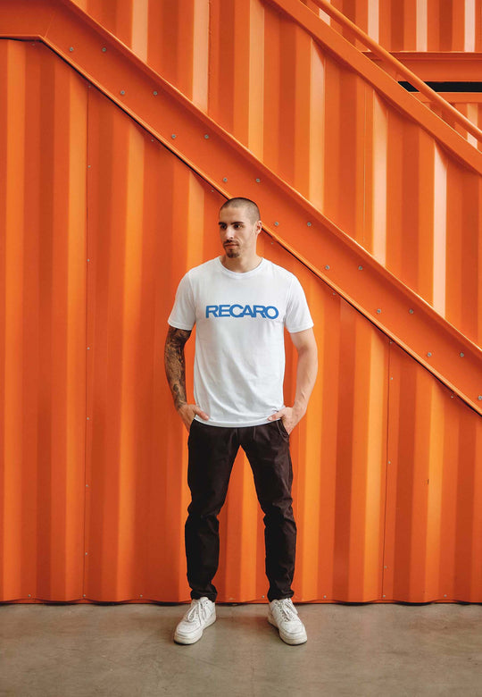 Vor einer orangefarbenen Metallwand und einer Treppe steht ein Mann mit kurzen Haaren und Tattoos. Er trägt ein T-Shirt Originals RECARO von Recaro_Merch_Daniel, eine schwarze Hose und weiße Sneakers und blickt zur Seite.