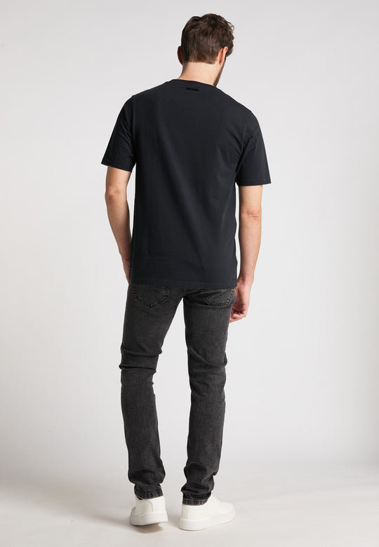 Ein Mann, von hinten gesehen, trägt das Recaro T-Shirt Originals Basic in schlichtem Schwarz mit entspannter Passform, gestylt mit dunkler Jeans und weißen Sneakers auf hellgrauem Untergrund.