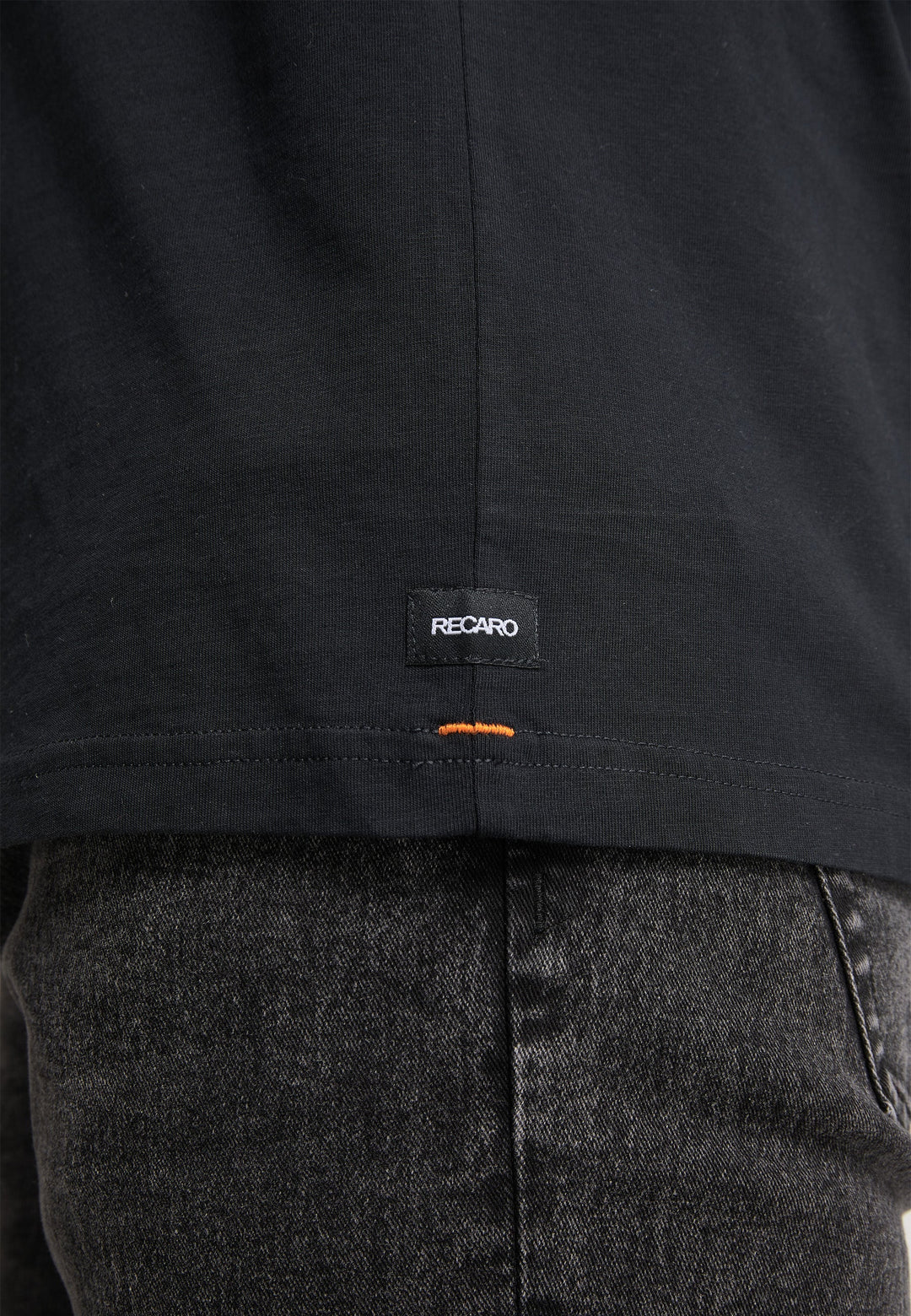 Nahaufnahme einer Person in dunkelgrauen Jeans und einem schwarzen Recaro_Merch_Daniel T-Shirt Originals Basic, das ein kleines schwarzes RECARO-Etikett mit einem orangefarbenen Stichdetail in der Nähe des Saums für einen entspannten Look zeigt.