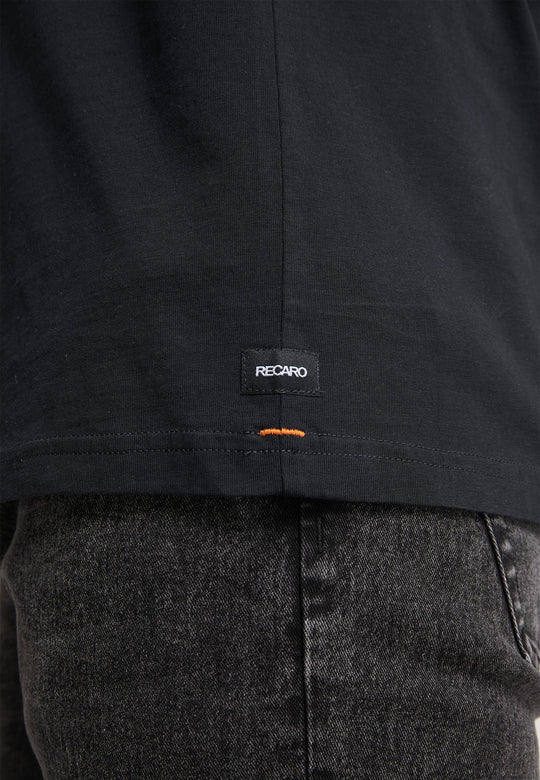 Nahaufnahme einer Person in dunkelgrauen Jeans und einem schwarzen Recaro_Merch_Daniel T-Shirt Originals Basic, das ein kleines schwarzes RECARO-Etikett mit einem orangefarbenen Stichdetail in der Nähe des Saums für einen entspannten Look zeigt.