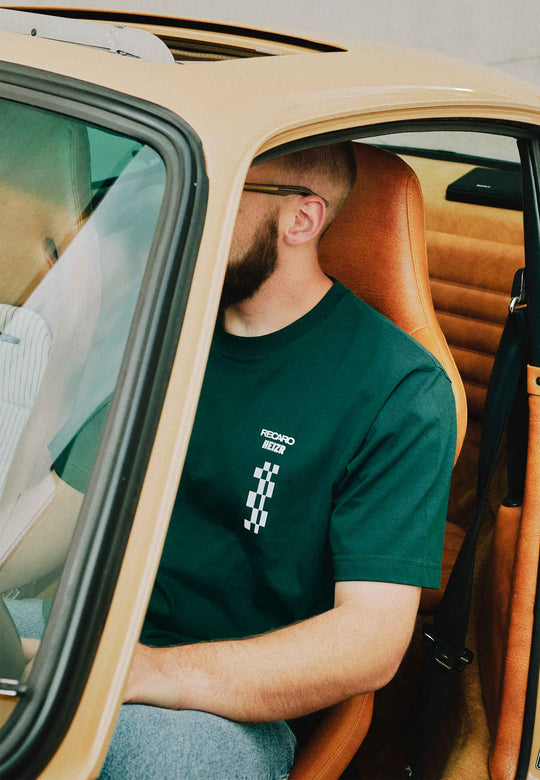 Ein bärtiger Mann mit Brille sitzt im hellbraunen Innenraum eines Oldtimers und trägt das T-Shirt HEIZR Club X RECARO aus dem RECARO Shop – ein klassischer Look für jedes HEIZR Club-Mitglied.