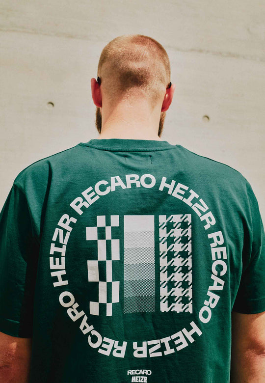 Von hinten gesehen trägt eine kurzhaarige, bärtige Person das grüne RECARO Shop T-Shirt HEIZR Club X RECARO mit kreisförmigem schwarz-weißem Karomuster und RECARO HEIZER-Design, inspiriert vom HEIZR Club-Style von Max Salzborn.