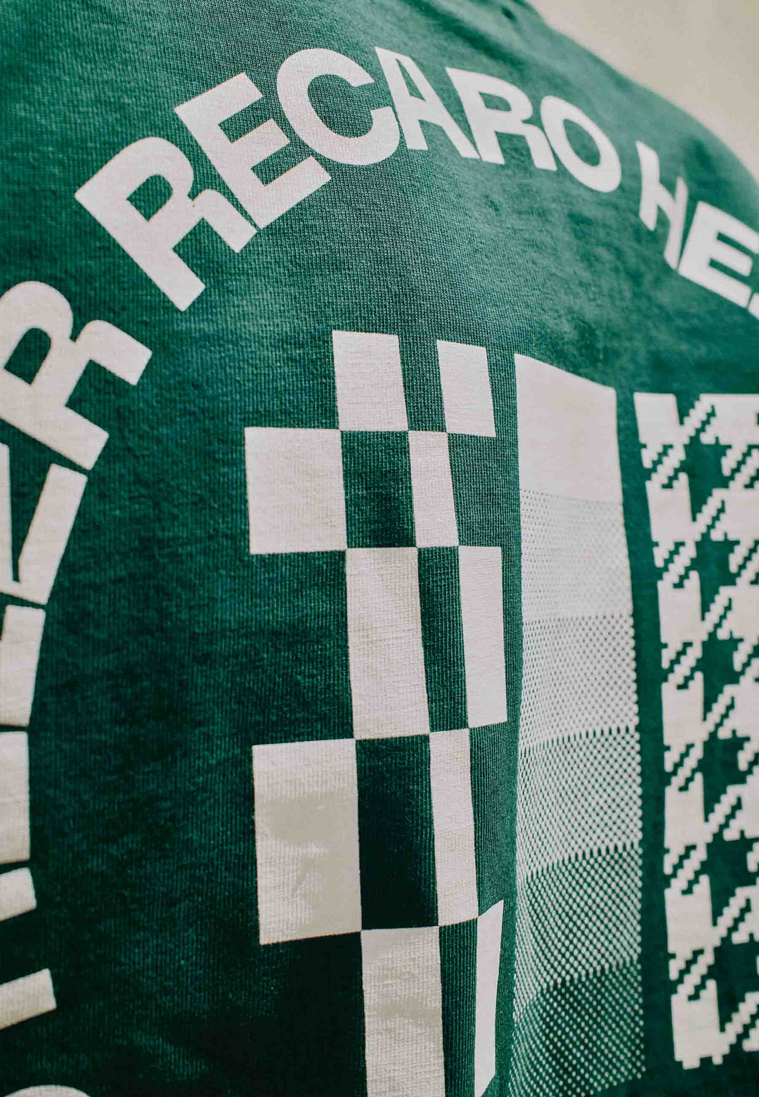 Nahaufnahme des grünen „T-Shirt HEIZR Club X RECARO“ aus dem RECARO Shop mit kräftiger weiß karierter Grafik und geschwungenem „RECARO“-Text – ein einzigartiges Kooperationsdesign.