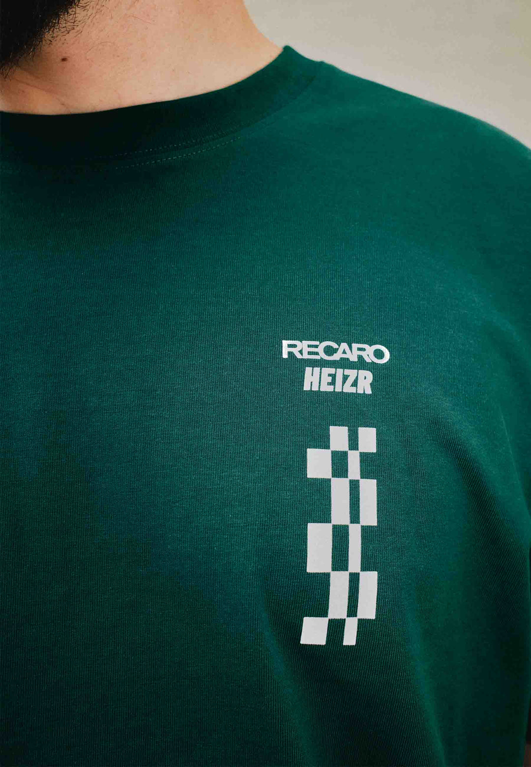 Nahaufnahme einer Person, die das T-Shirt HEIZR Club X RECARO aus dem RECARO Shop trägt. Auf dem T-Shirt steht „RECARO HEIZR“ in Weiß über einer vertikalen Zielflagge – ein charakteristischer Stil, der von HEIZR Club-Fans und Max Salzborn-Enthusiasten geliebt wird.