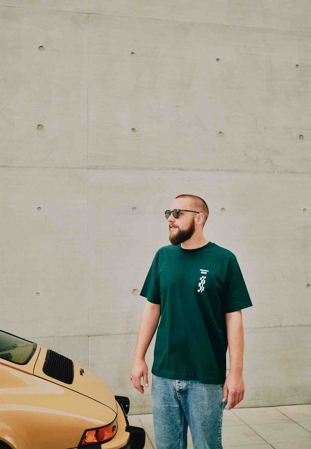 Ein bärtiger Mann mit Sonnenbrille, Jeans und grünem T-Shirt HEIZR Club X RECARO aus dem RECARO Shop steht im Freien neben einem gelben Oldtimer, im Hintergrund eine hellgraue Betonwand.