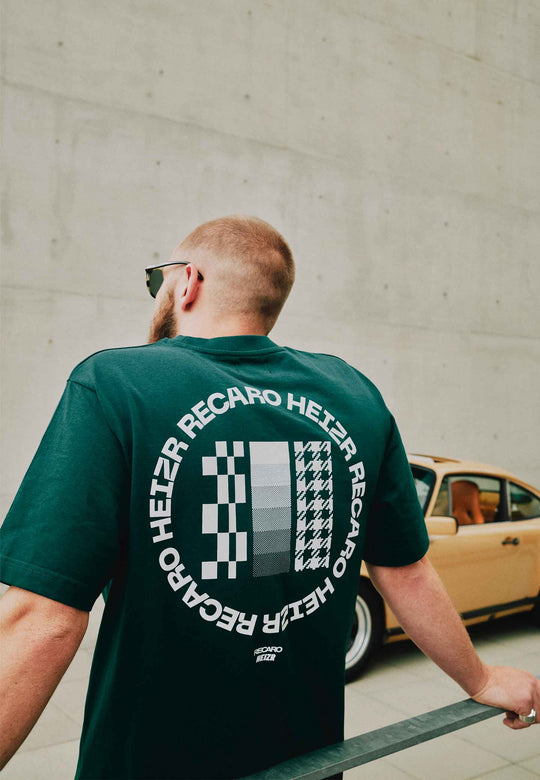 Eine Person mit kurzen Haaren und Sonnenbrille lehnt an einem Geländer. Sie trägt das T-Shirt „HEIZR Club X RECARO“ aus dem RECARO Shop. Auf dem Rücken ist „HEIZR RECARO“ aufgedruckt. Im Hintergrund ist ein gelbes Oldtimerauto aus der HEIZR Club-Szene zu sehen.