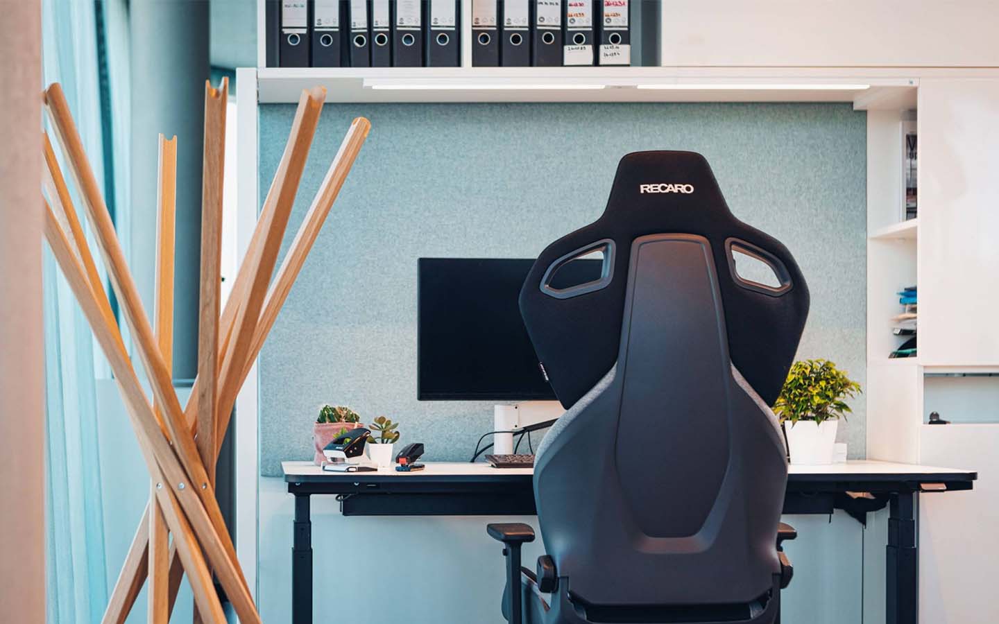 Ein moderner Büroarbeitsplatz mit einem schwarzen ergonomischen Stuhl am Schreibtisch, einem Computermonitor, Pflanzen und darüber Regalen mit Ordnern. Links steht eine hölzerne Garderobe. Der Raum ist ordentlich und gut organisiert.