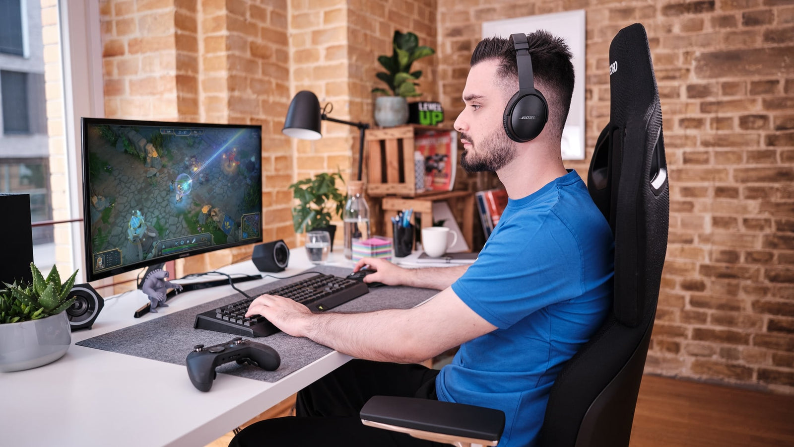 Ein Mann mit Kopfhörern sitzt an einem Schreibtisch und spielt ein Spiel auf einem Computermonitor. Auf dem Schreibtisch befinden sich Tastatur, Maus, Gamecontroller und Pflanzen. Der Raum hat Ziegelwände, Regale und ein Fenster.