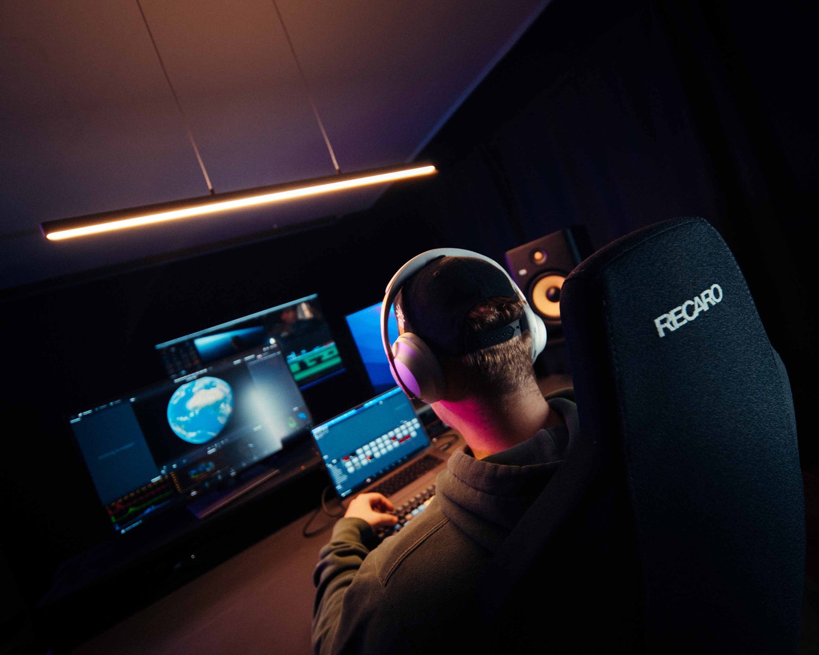 Eine Person mit Kopfhörern sitzt in einem dunklen Raum an einem Schreibtisch. Auf mehreren Monitoren werden Bearbeitungssoftware und ein Globusbild angezeigt. Zum Setup gehören ein Lautsprecher und ein RECARO-Stuhl.